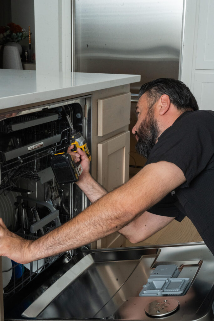 Technician repairing a dishwasher with a power tool in a modern kitchen