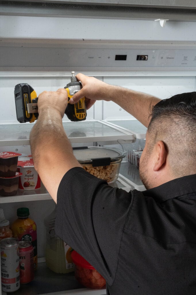 Person using a power drill to adjust shelves inside a refrigerator