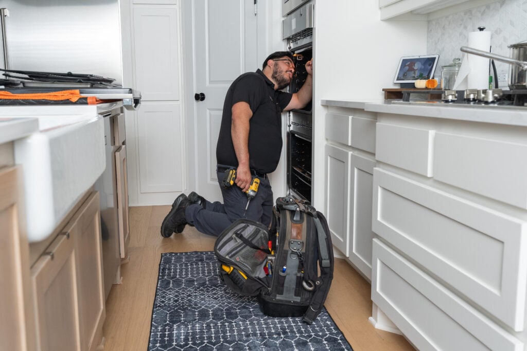 Technician kneeling in kitchen, inspecting oven with tools and gear