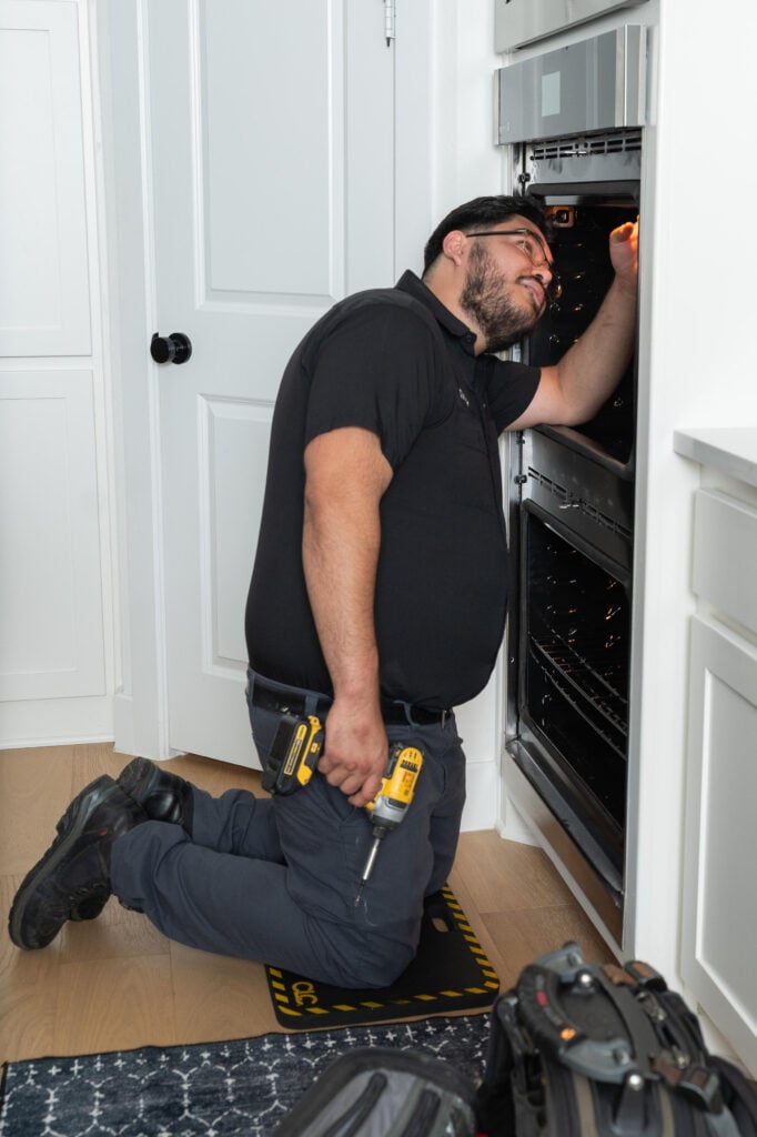 Technician kneeling by kitchen oven, using a drill for repairs
