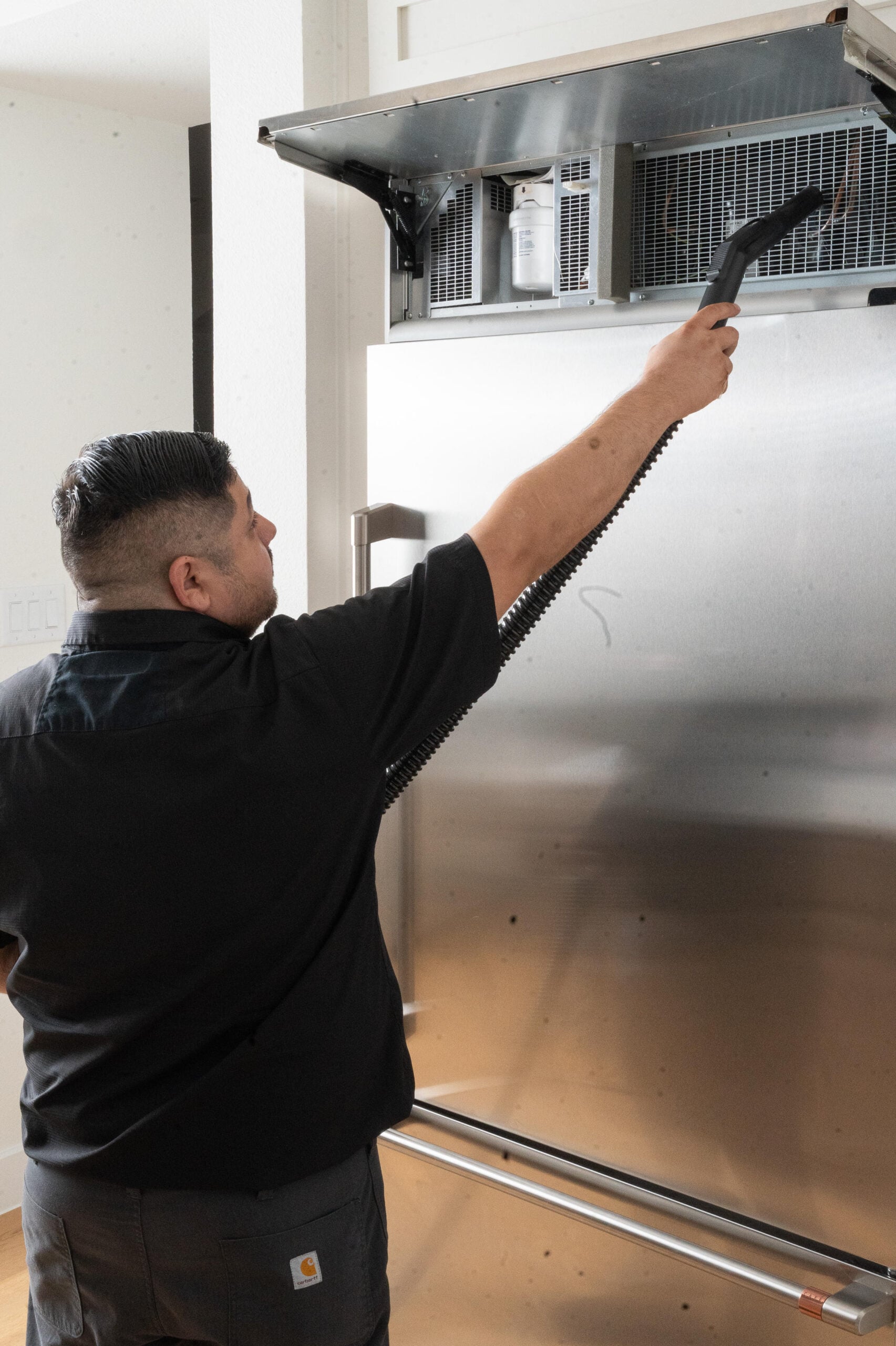 Man cleaning the top of a stainless steel refrigerator with a handheld vacuum