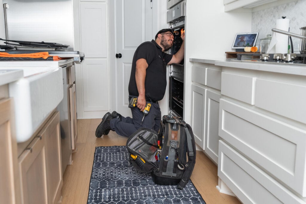 A technician kneeling in a kitchen, examining an oven with a toolkit beside him